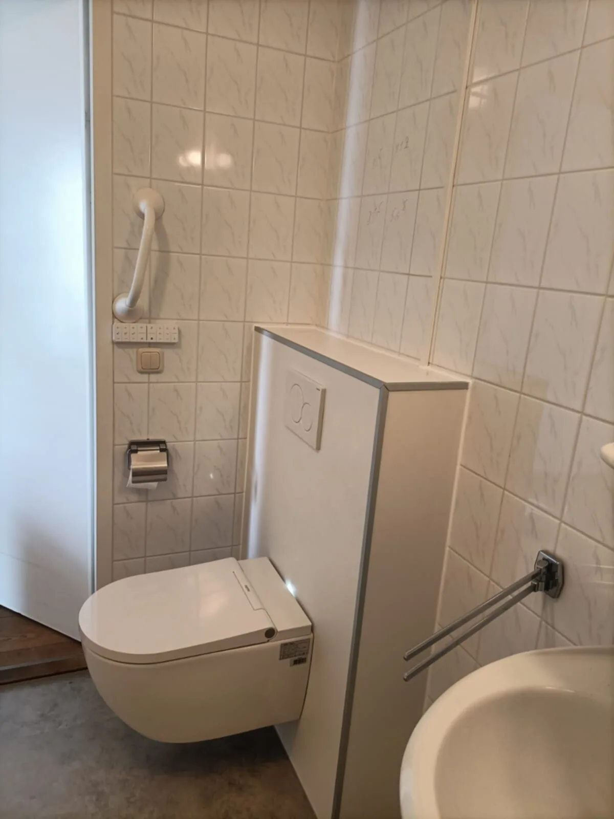 Hangend douchetoilet tegen een witte voorzetwand met steunbeugel en tegels