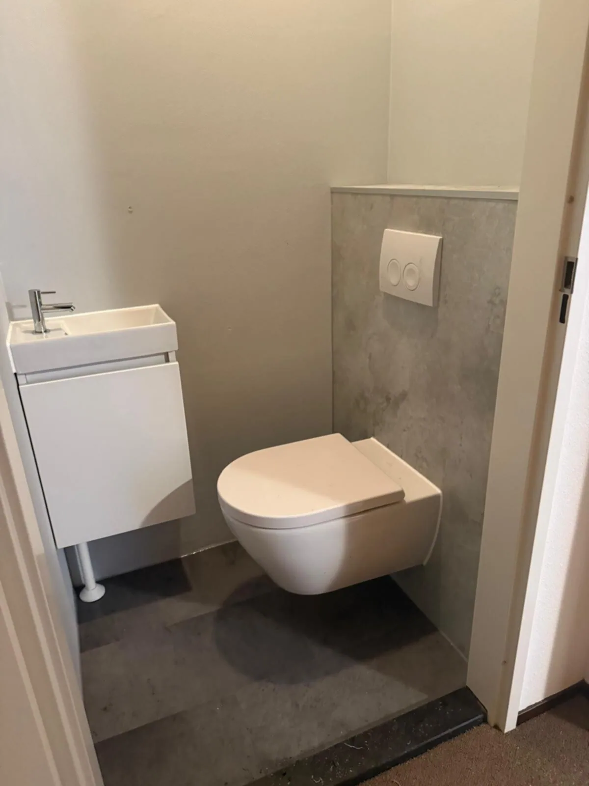 Hangtoilet tegen een betonlook voorzetwand met witte drukplaat en een wit fonteinmeubeltje ernaast