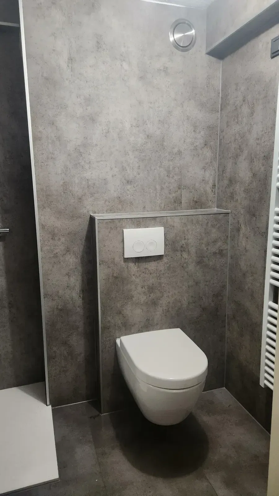 Wit hangtoilet met drukplaat op een voorzetwand, wanden in grijze betonlook panelen