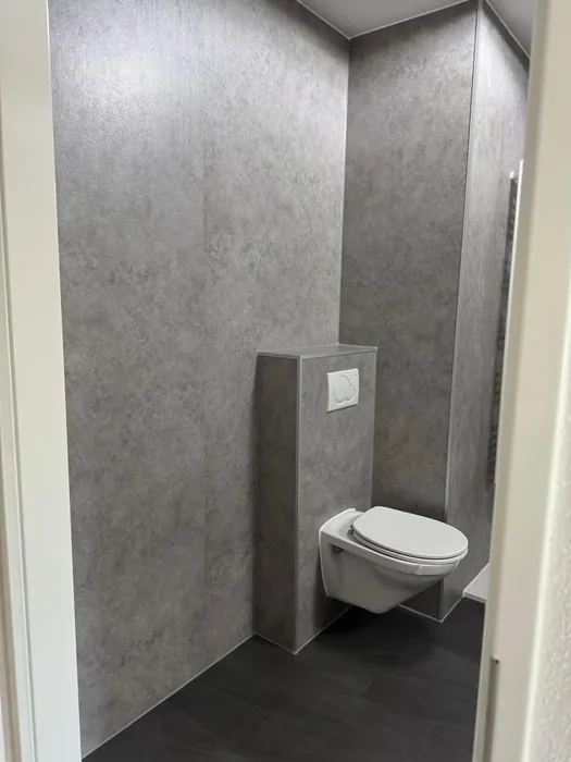 Hangtoilet tegen een voorzetwand, de hele ruimte bekleed met grijze steenlook panelen