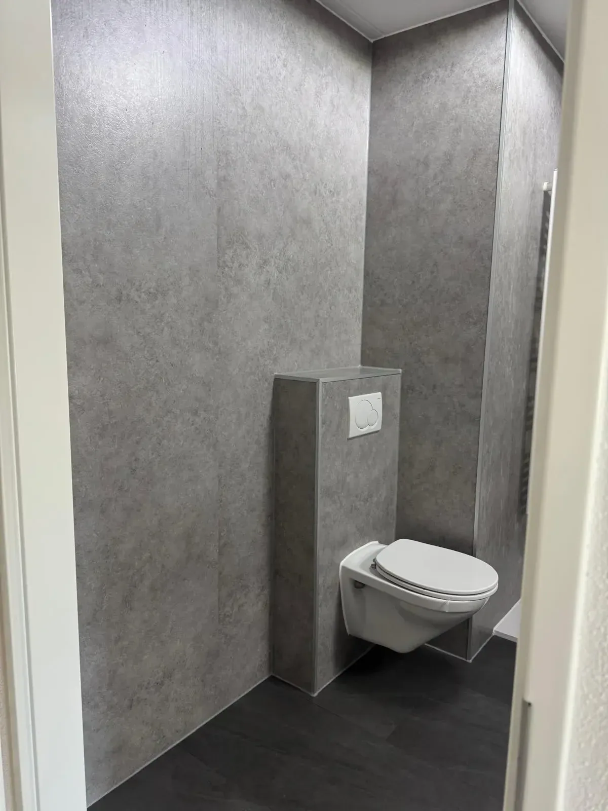 Hangtoilet tegen een voorzetwand, de hele ruimte bekleed met grijze steenlook panelen