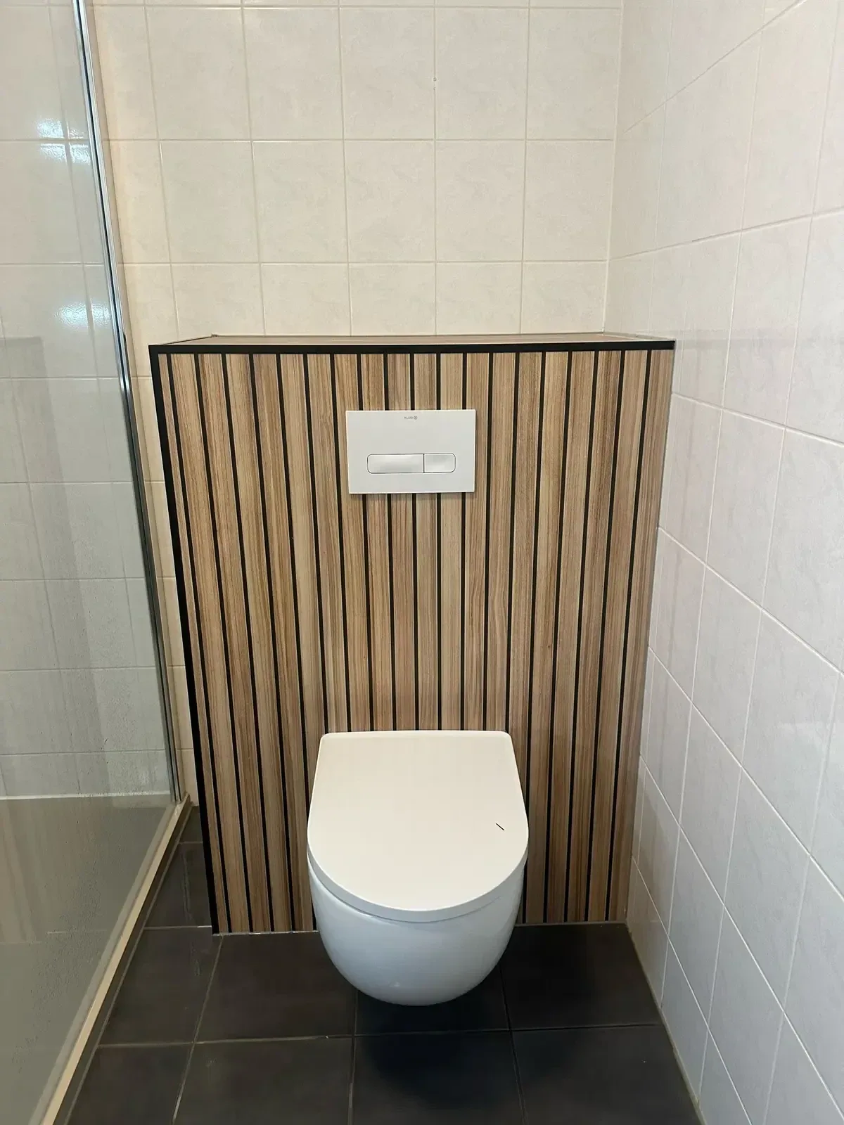Wit hangtoilet op een voorzetwand bekleed met houtlook lamellen, met witte tegels en donkere vloer