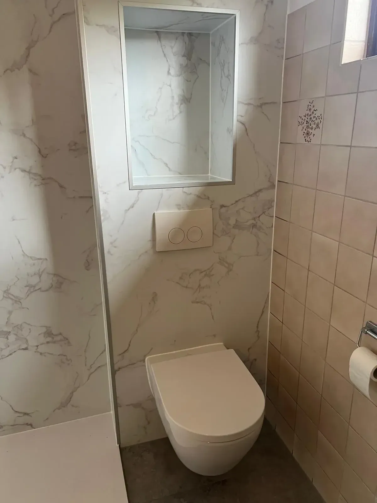Hangtoilet tegen een marmerlook paneel met drukplaat en nis, naast bestaande beige tegels