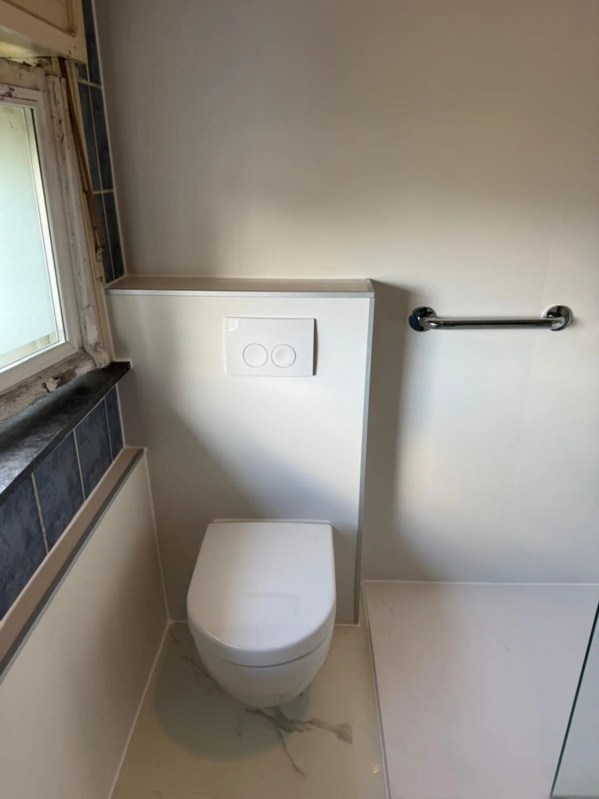 Hangtoilet tegen een witte voorzetwand met drukplaat en steunbeugel, op een vloer in marmerlook