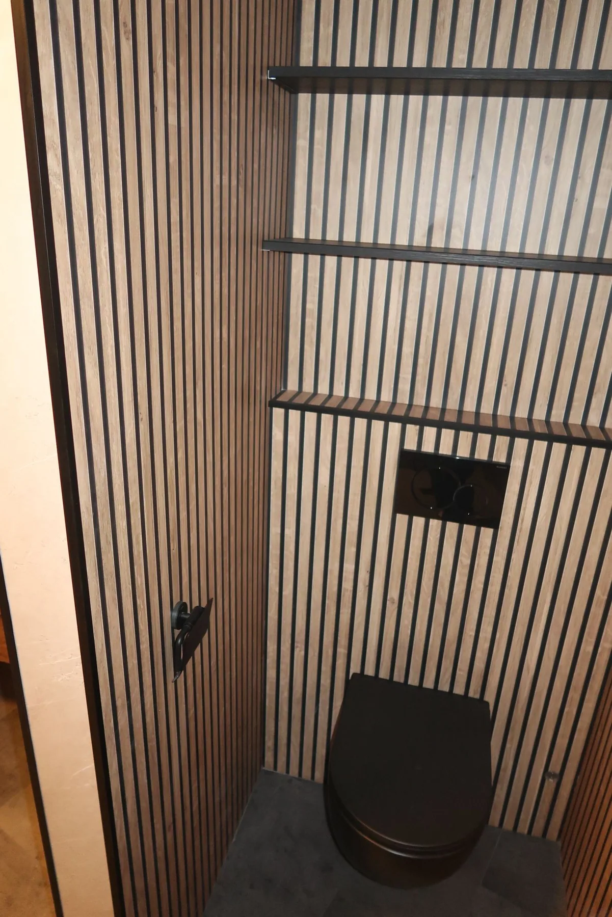 Drie zwarte wandplanken boven een zwart hangtoilet tegen een houten lamellenwand
