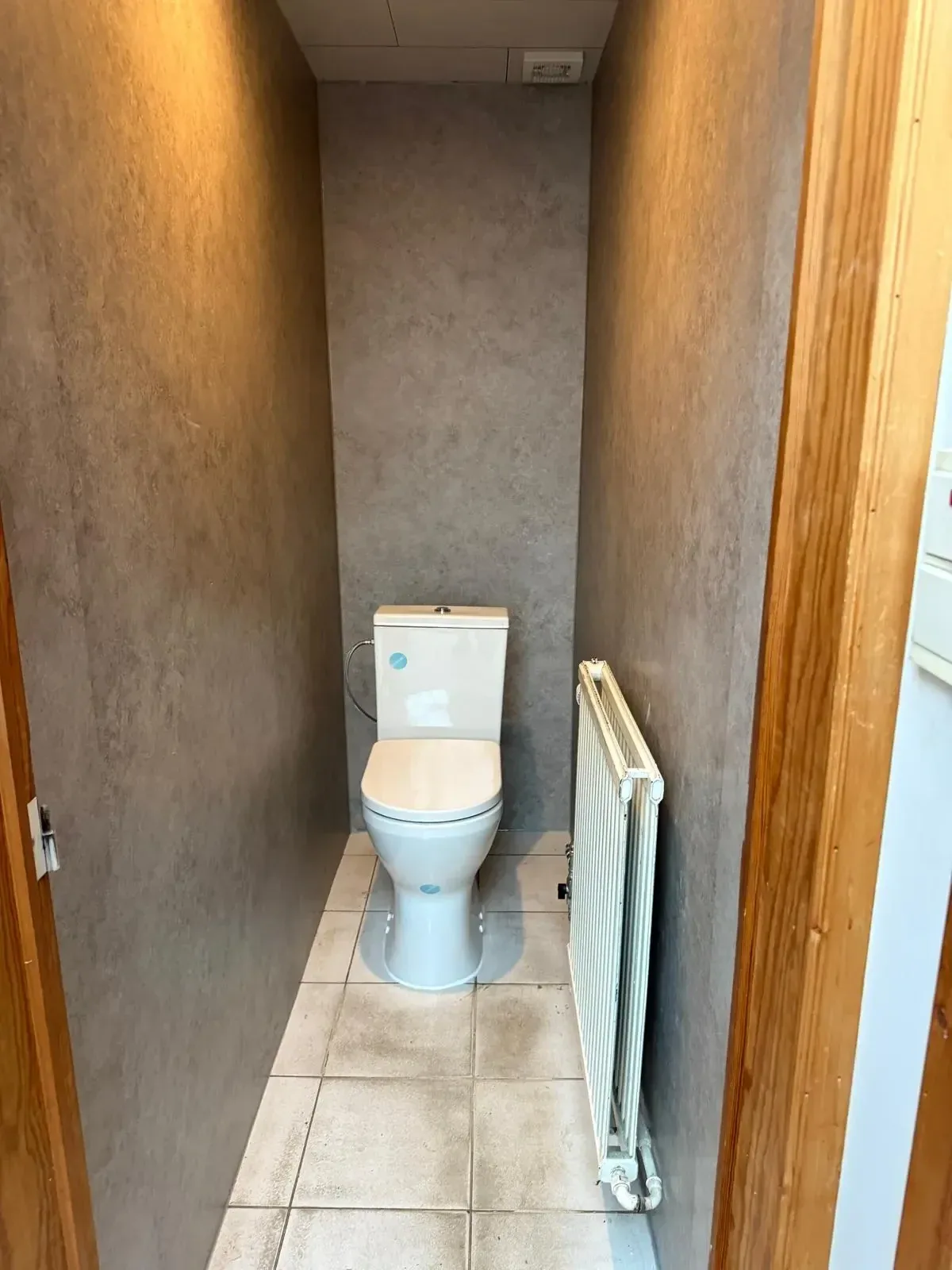 Smalle wc met betonlook wandpanelen, een staand toilet en een radiator