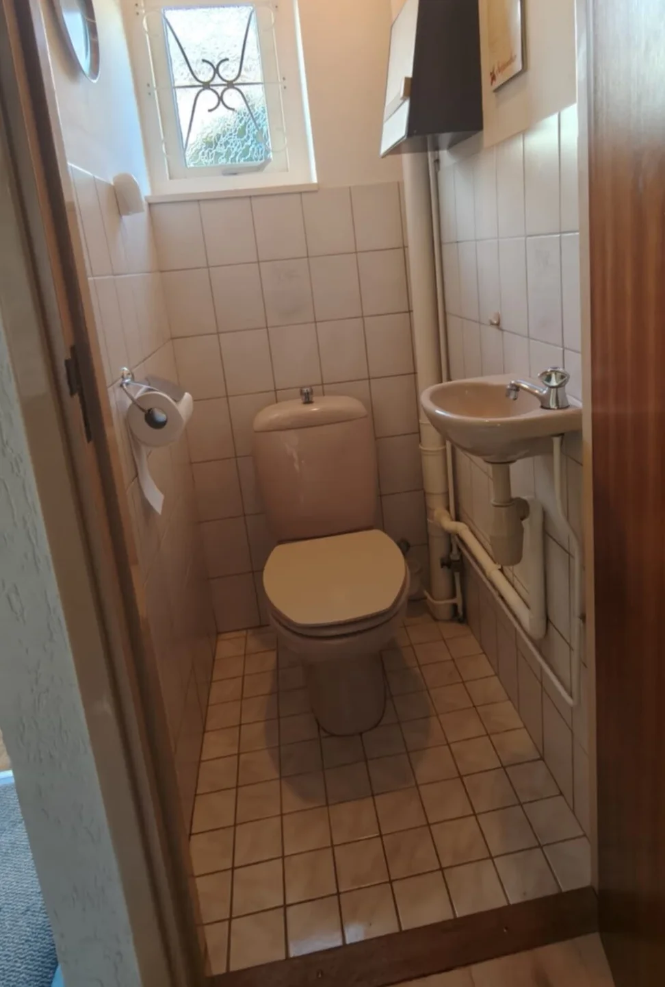 Vóór: smalle wc met beige staand toilet, klein handwasbakje en witte tegels onder een raampje met sierrooster