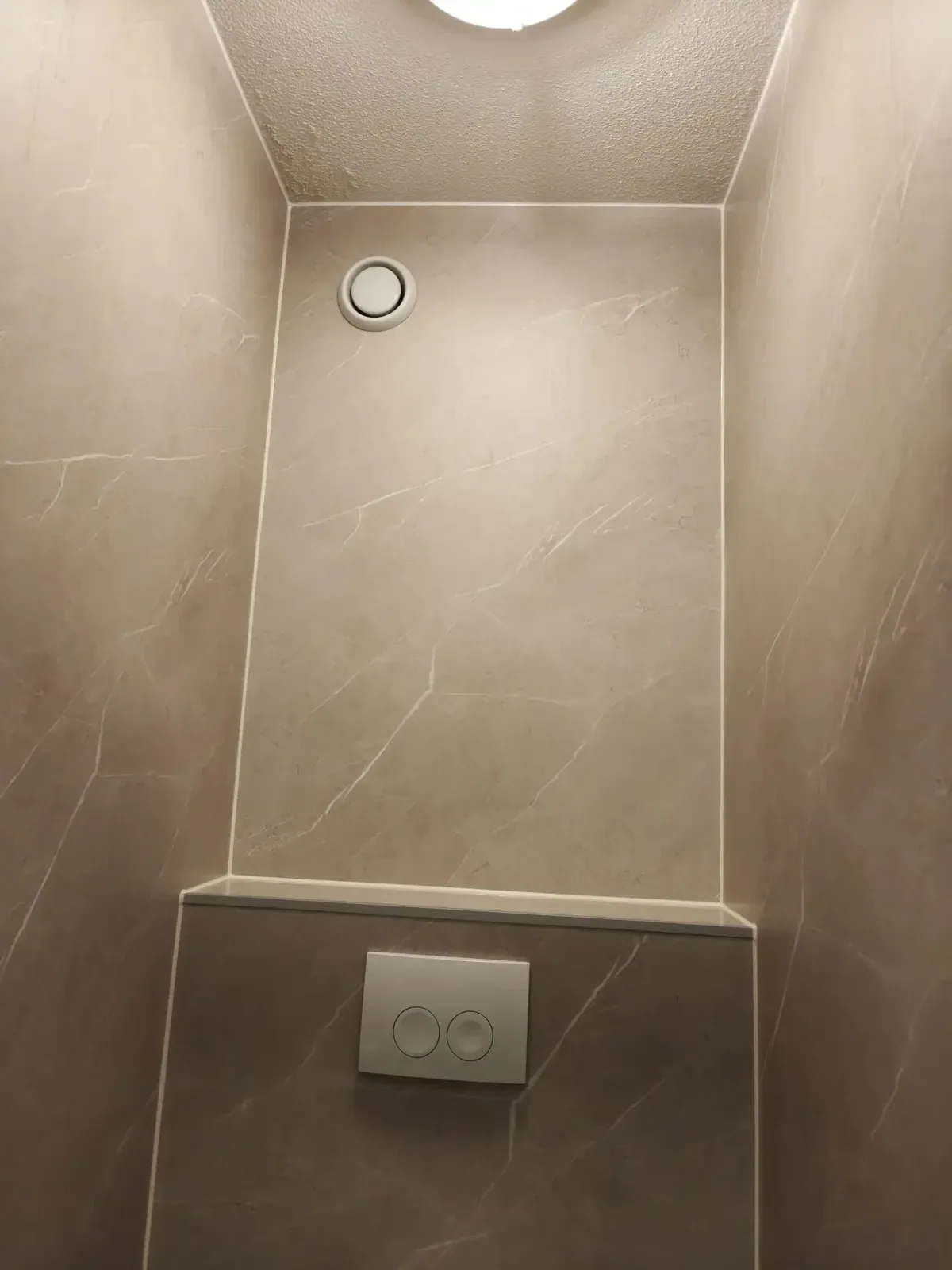 Wc met beige marmerlook panelen, voorzetwand met drukplaat en een ventilatierooster in het plafond