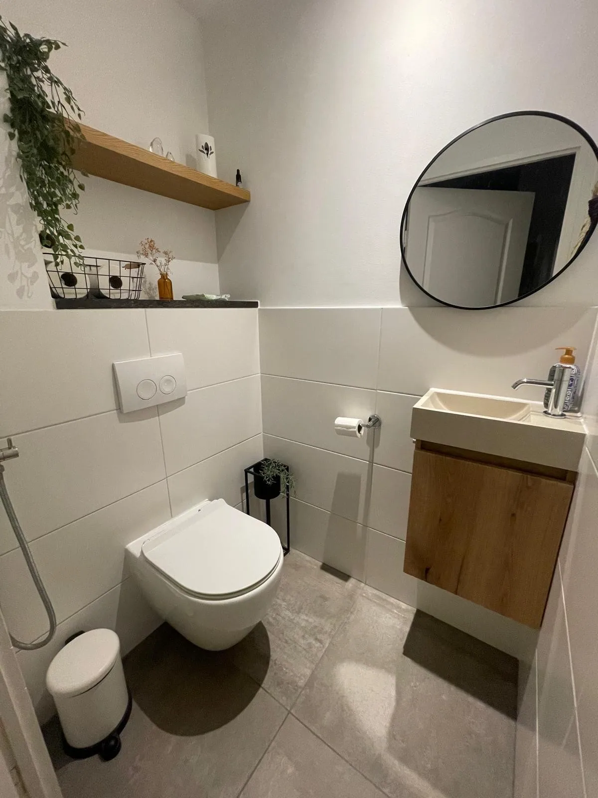 Wc met hangtoilet, houten fonteinkastje, ronde zwarte spiegel en grote grijze vloertegels