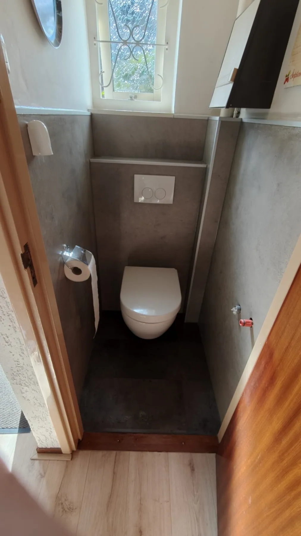 Na de renovatie: hangtoilet, grijze betonlook wandpanelen en voorzetwand met drukplaat onder het raampje