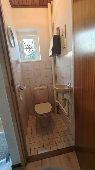 Vóór de renovatie: smalle wc met beige staand closet, handwasbakje, witte tegels en een raampje met sierrooster